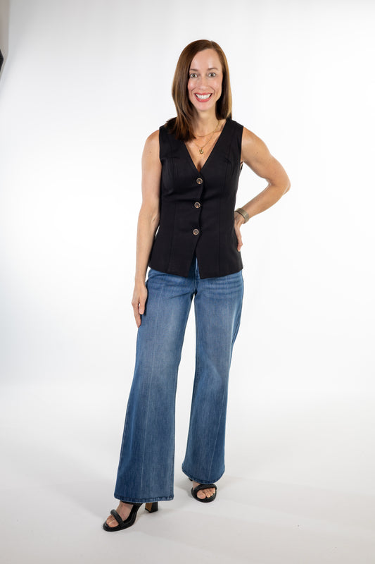 A woman modeling a sleeveless black vest with three front buttons and wide-leg blue jeans against a plain white studio background.
