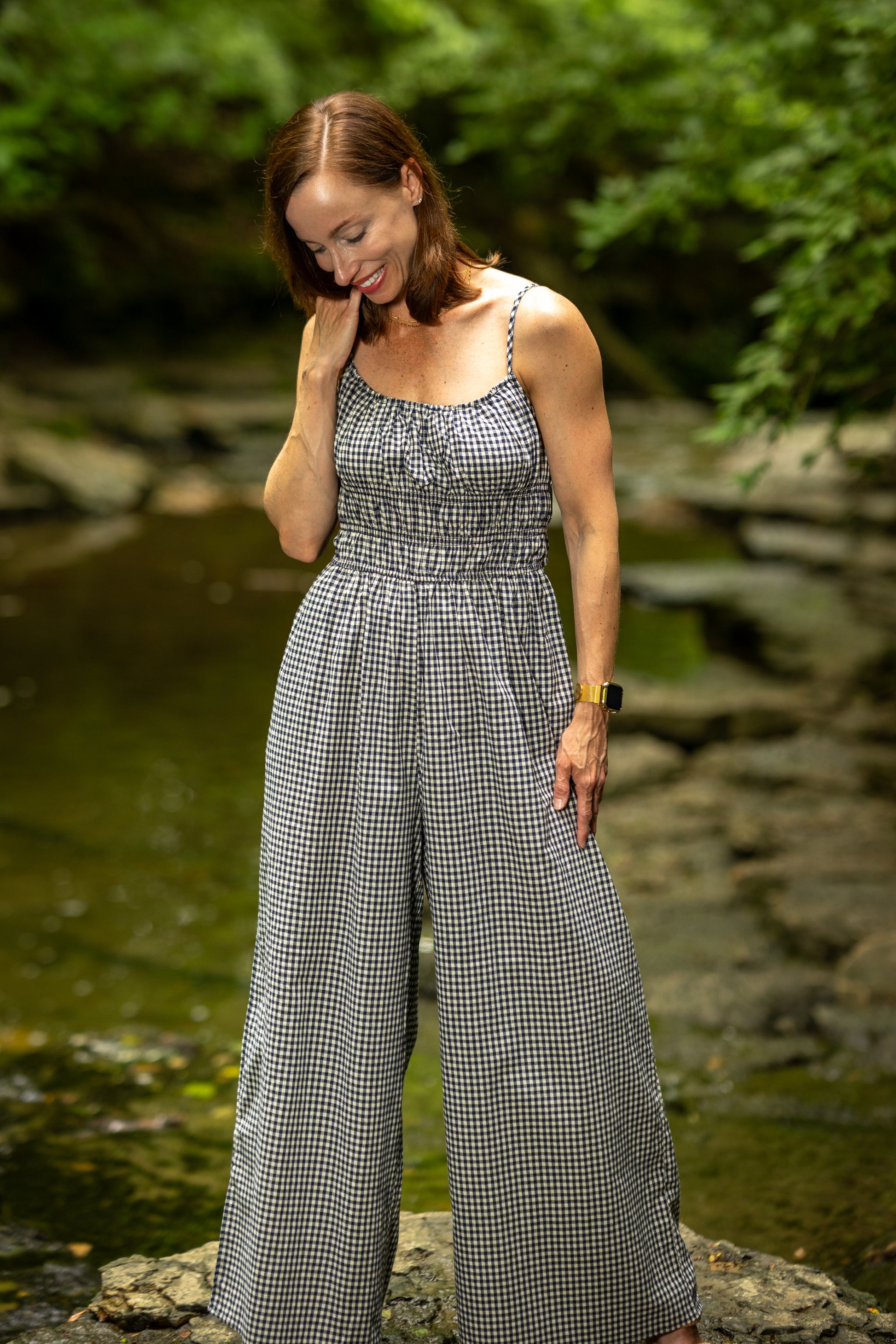 Woman in a checkered jumpsuit standing by a stream with greenery in the background