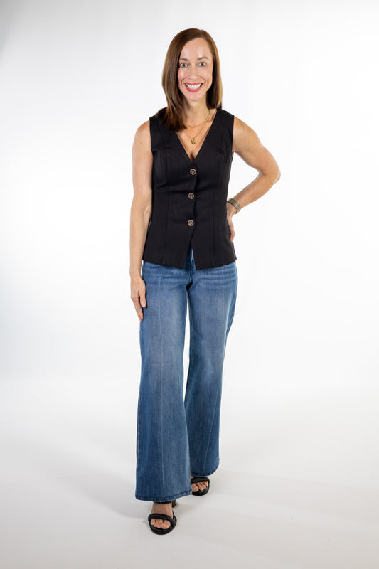 A woman modeling a sleeveless black vest with front buttons, paired with blue wide-leg jeans and black sandals, standing against a white studio background.