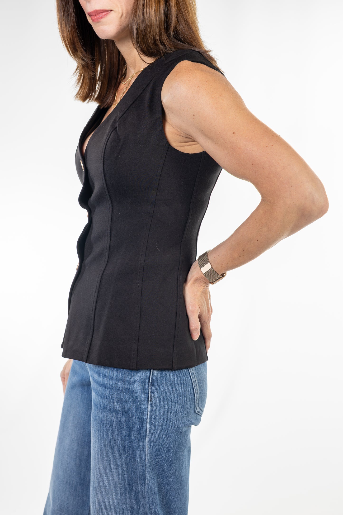 A woman wearing a black sleeveless wrap-style vest top with structured seams, paired with blue jeans, shown from the side against a plain white studio background.