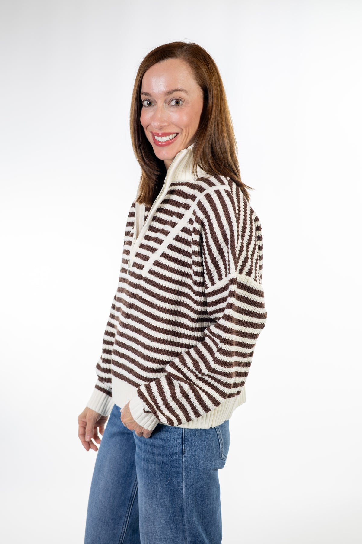 A woman wearing a brown and cream striped knit sweater with a half-zip neckline, paired with blue jeans, photographed against a solid white studio background.