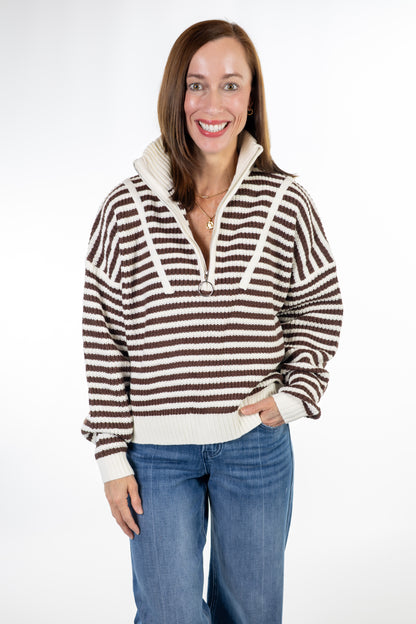 A woman wearing a brown and cream striped knit half-zip sweater with a high collar, paired with blue jeans, photographed against a plain white studio background.