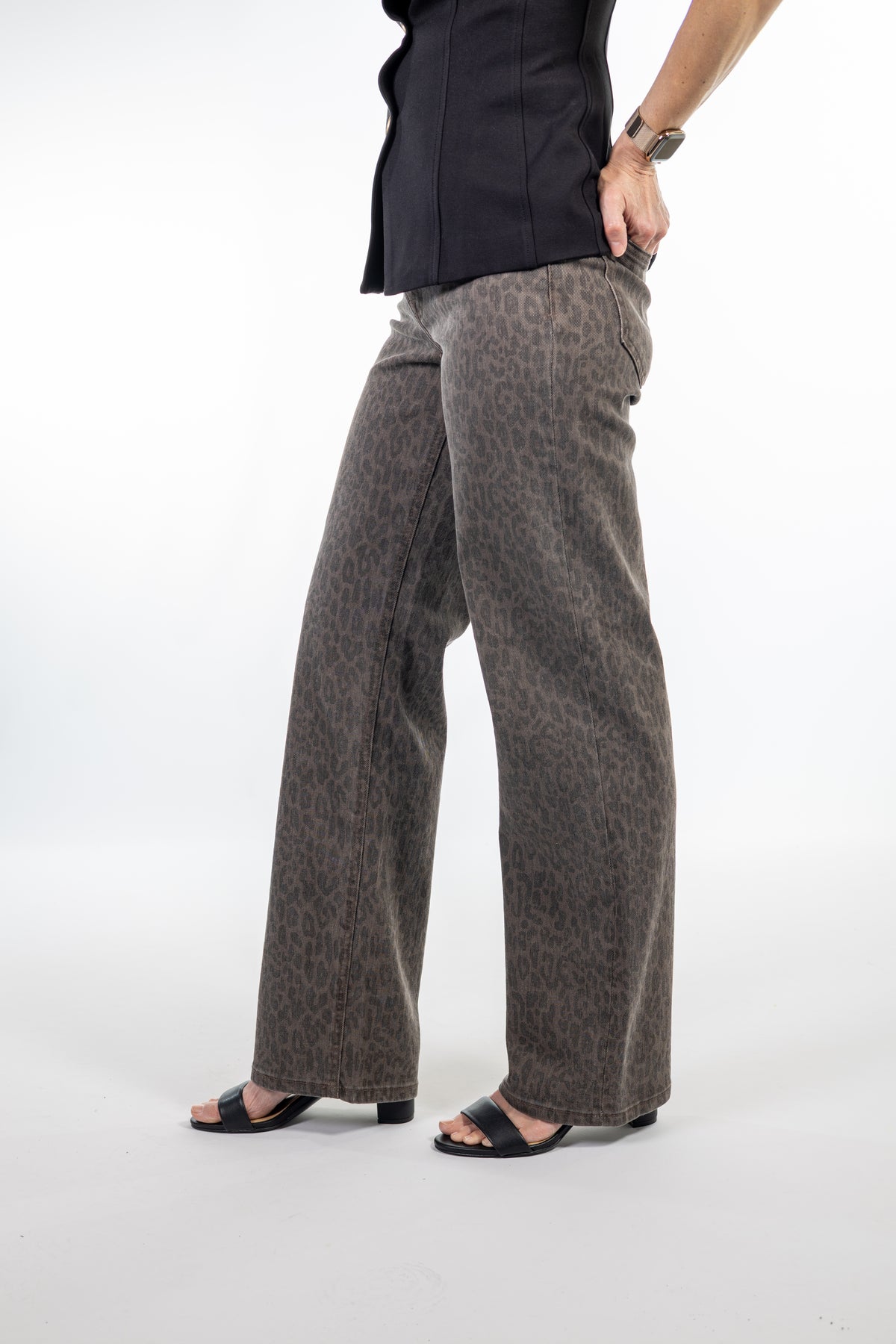 A person wearing wide-leg brown and black leopard-print pants, paired with a black top and black slide sandals, standing against a white studio background.
