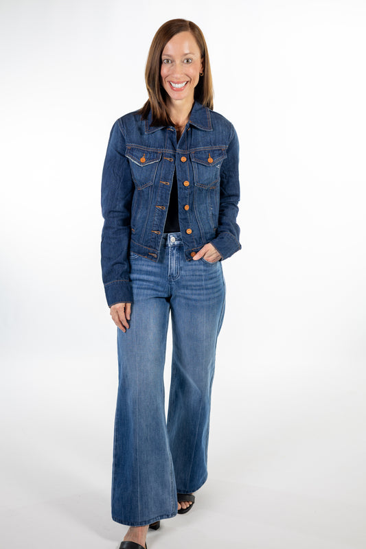 A woman modeling a dark wash blue denim jacket and matching wide-leg jeans with copper button accents, standing against a plain white studio background.