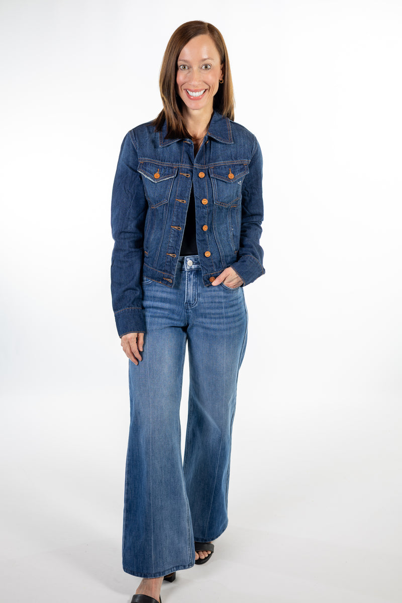 A woman modeling a dark wash blue denim jacket and matching wide-leg jeans with copper button accents, standing against a plain white studio background.