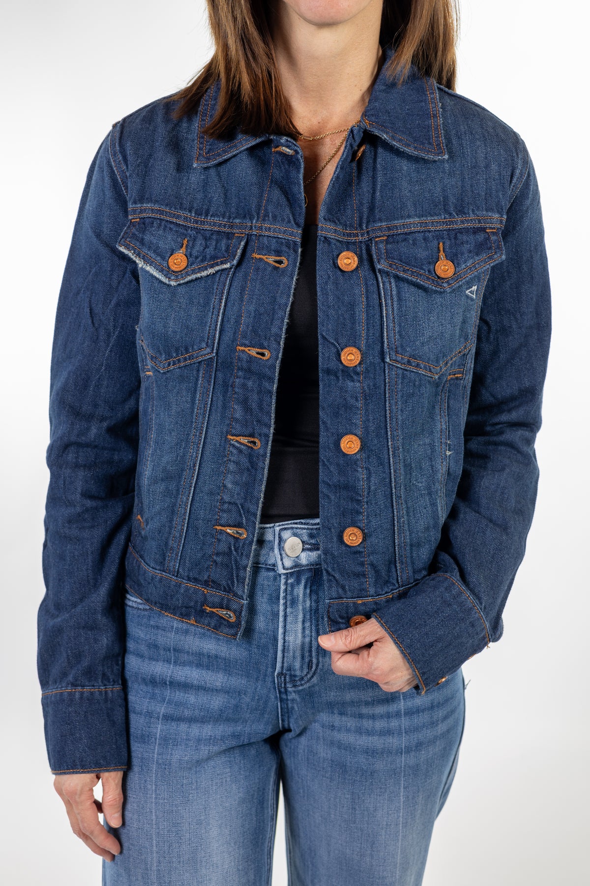 A woman wearing a cropped dark blue denim jacket with copper buttons, styled with blue jeans and a black top, shown from the neck down against a light gray studio background.
