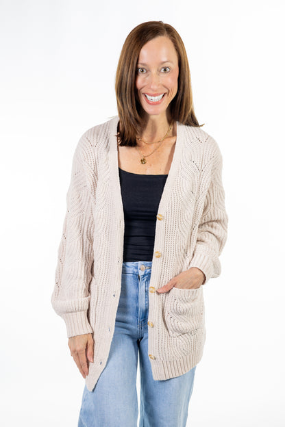 A woman modeling a cream-colored cable-knit cardigan with gold buttons over a black tank top and light blue jeans, photographed against a solid white studio background.