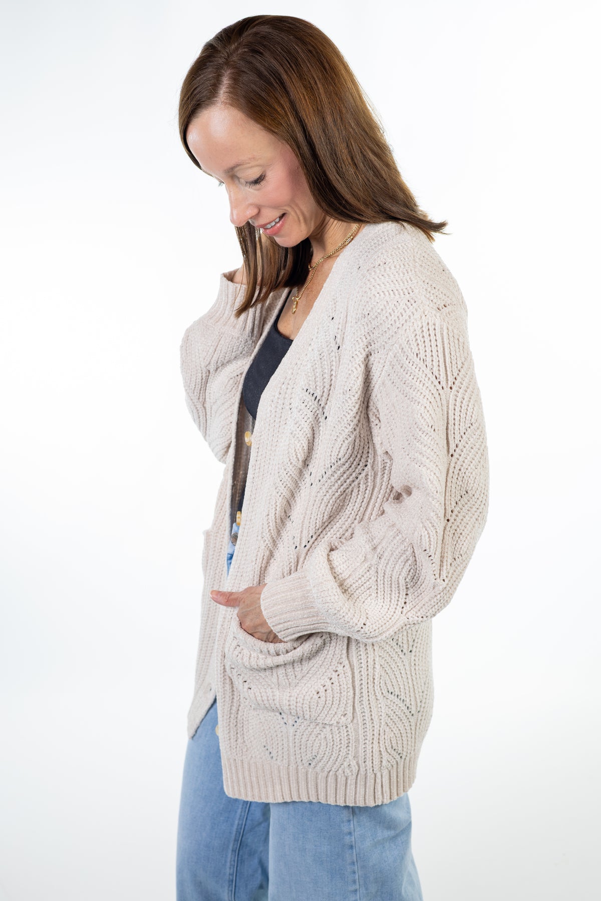 A woman wearing a cream cable-knit cardigan over a black top and blue jeans, standing against a plain white studio background.