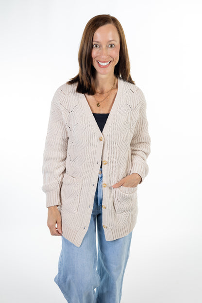 A woman modeling a cream cable-knit cardigan with gold buttons, worn over a black top and blue jeans, against a white studio background.