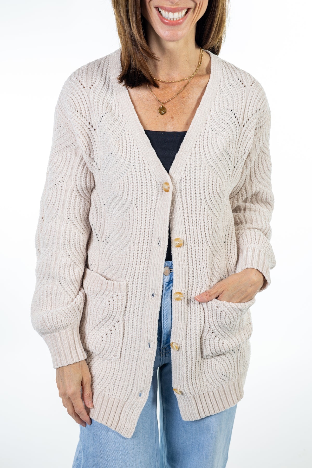 A woman wearing a cream cable-knit cardigan with gold buttons over a black top and blue jeans, shown from the waist up against a white background.