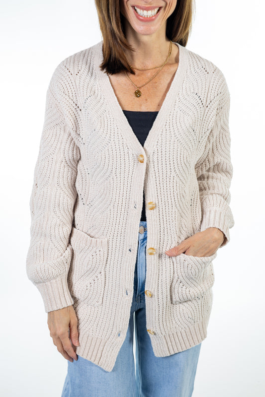 A woman wearing a cream cable-knit cardigan with gold buttons over a black top and blue jeans, shown from the waist up against a white background.