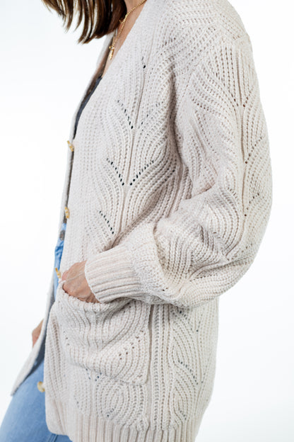 A close-up side view of a person wearing a chunky knit cardigan in a light oatmeal beige color, paired with blue jeans and a gold necklace against a plain white background.