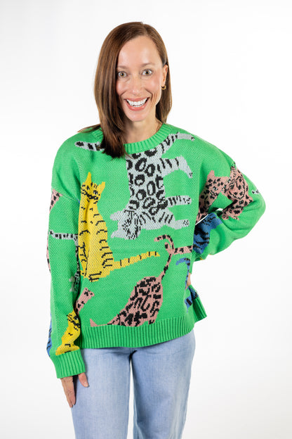 A woman wearing a bright green knit sweater with a colorful pattern of stylized tiger cats in yellow, pink, black, and gray, paired with light blue jeans, against a plain white background.