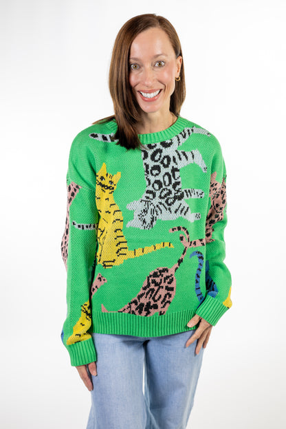 A woman wearing a bright green knit sweater with a colorful pattern of stylized cats in yellow, pink, black, and white, paired with light blue jeans, against a plain white background.