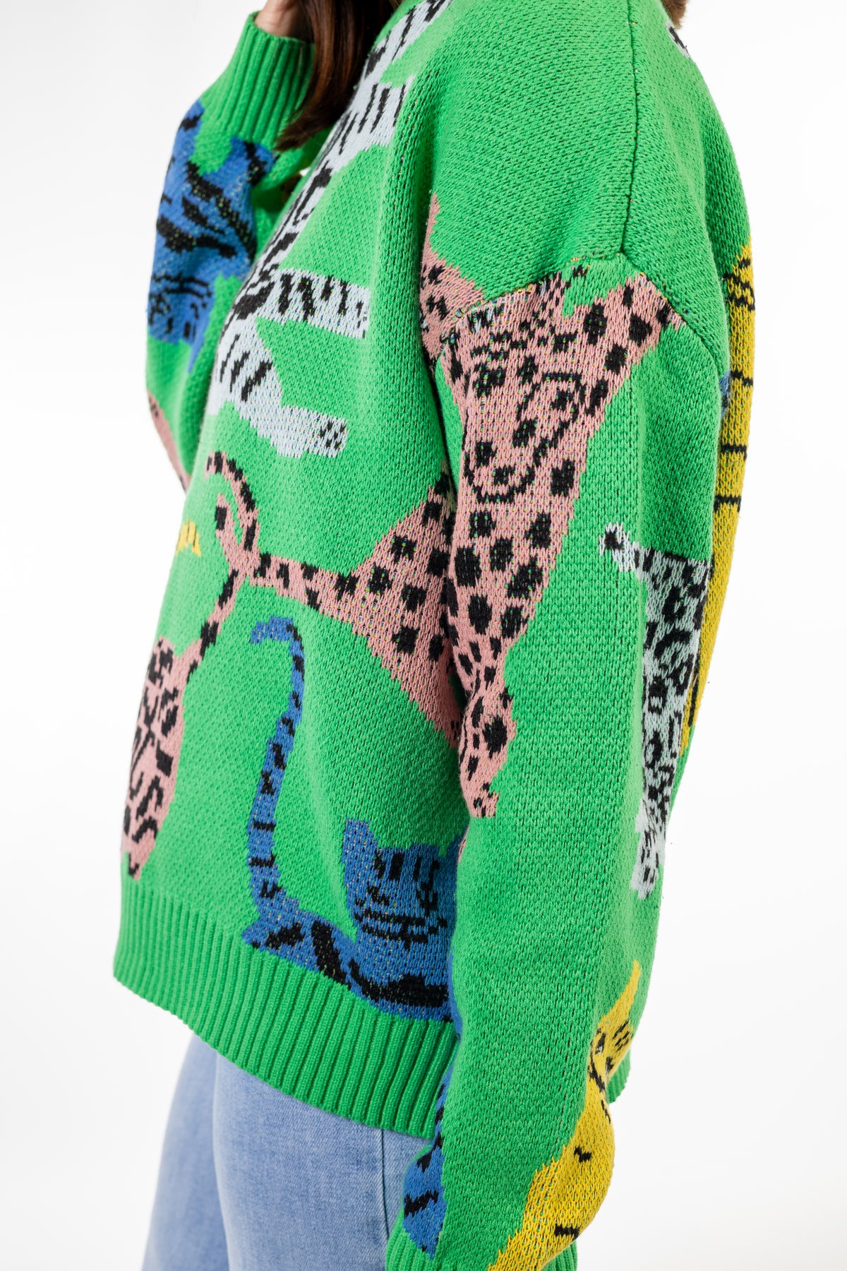 A close-up of a bright green knit sweater featuring a colorful animal  wild cat print pattern with pink, blue, yellow, and black-and-white spotted motifs, worn by a model with light blue jeans visible at the bottom.