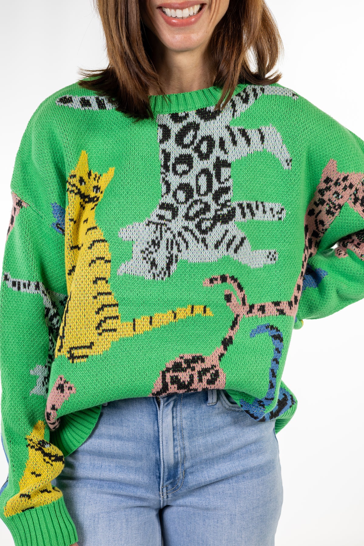 A woman wearing a bright green knit sweater with colorful animal cat tiger motifs, including a large black-and-white leopard print cat and yellow, pink, and blue spotted cats, paired with light blue jeans.