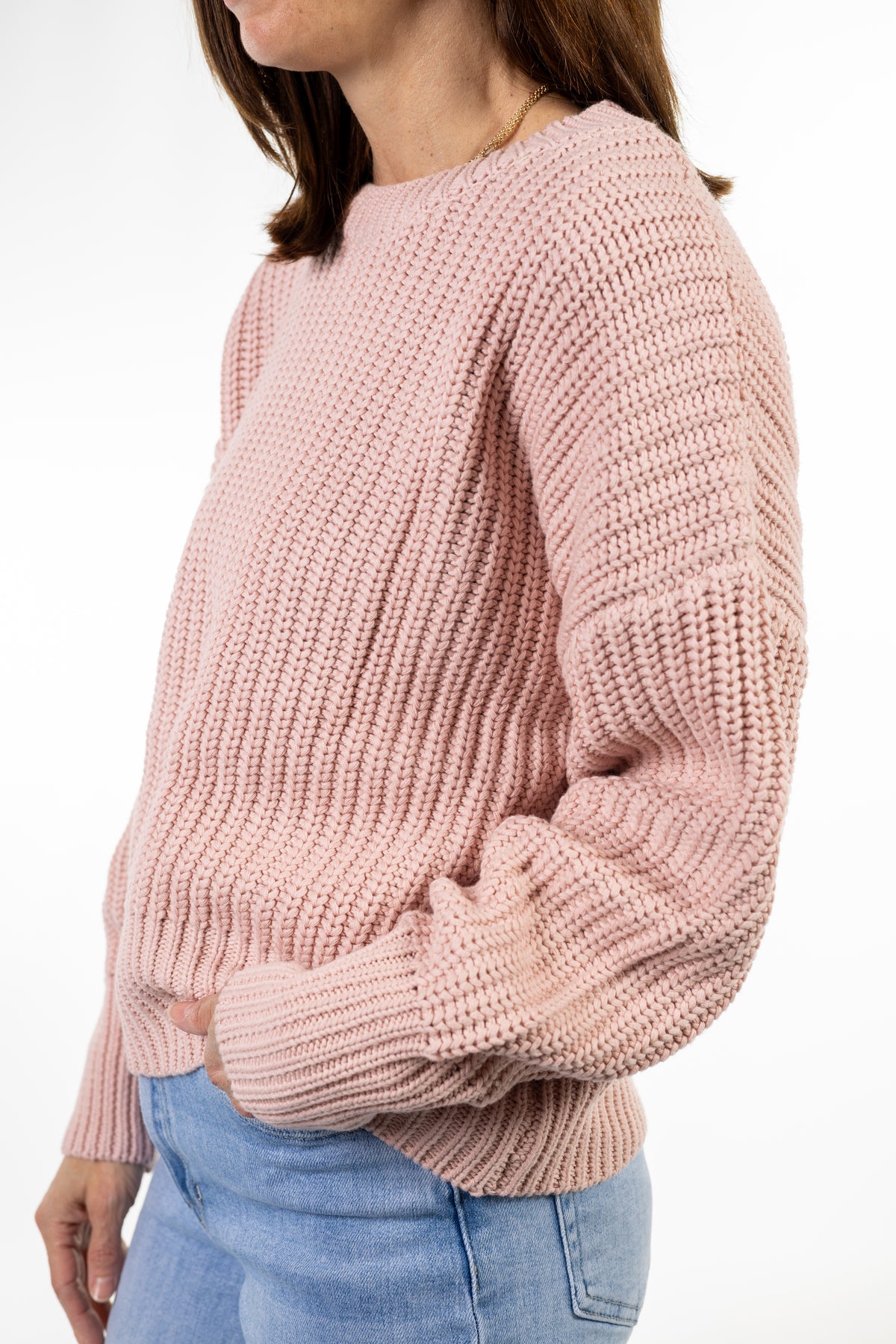 A woman wearing a dusty pink chunky knit sweater and light blue jeans, shown from the side against a plain white background.