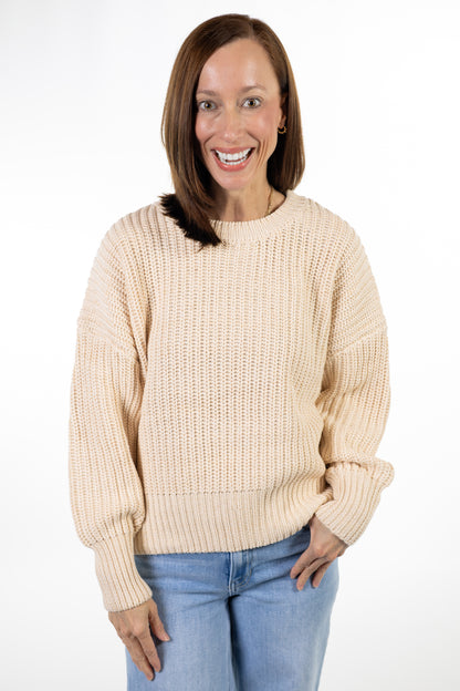 A smiling woman with shoulder-length brown hair wearing a cream-colored chunky knit sweater and light blue jeans, standing against a plain white studio background.