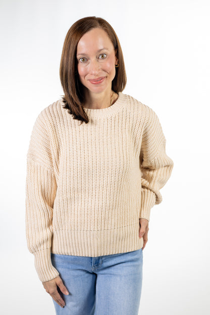 A woman with shoulder-length brown hair wearing a cream-colored ribbed knit sweater and light blue jeans, standing against a plain white studio background.