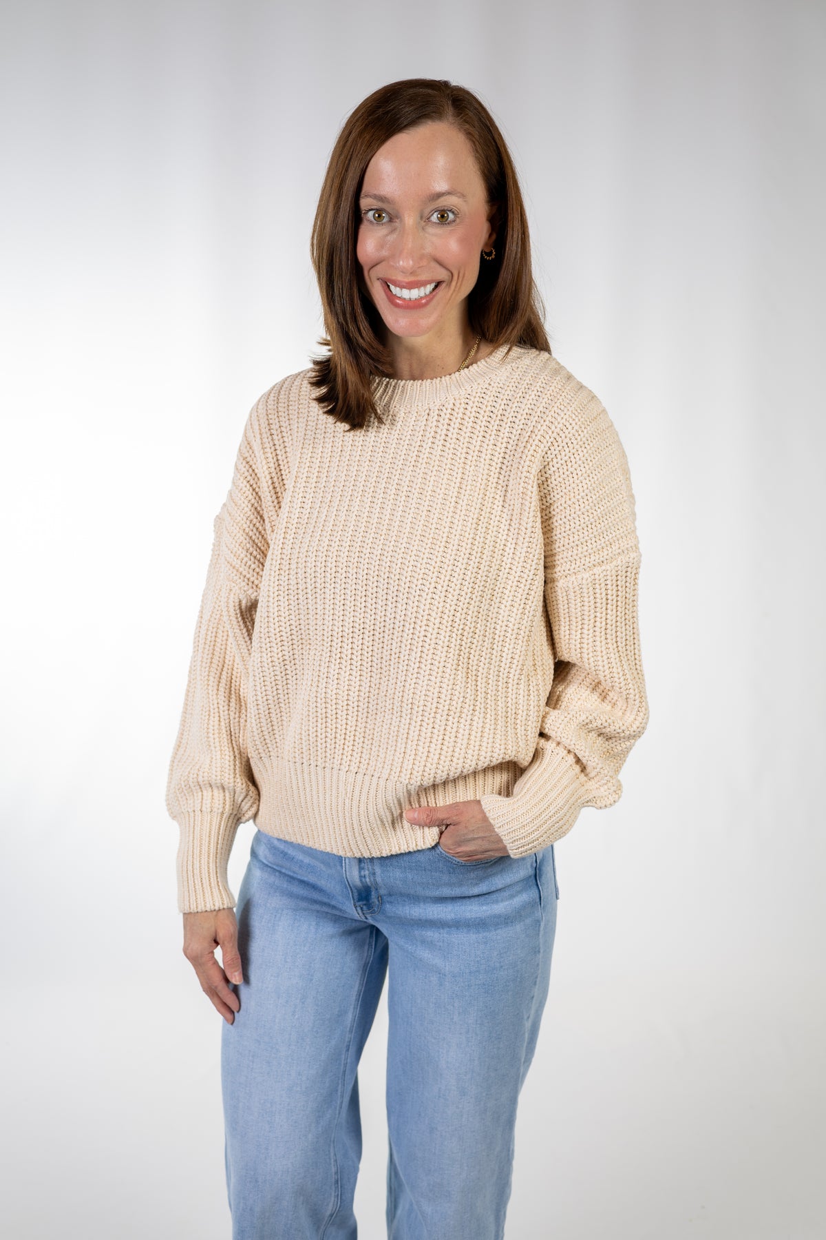 A smiling woman wearing a cream-colored chunky knit sweater and light blue jeans, standing against a light gray studio background.