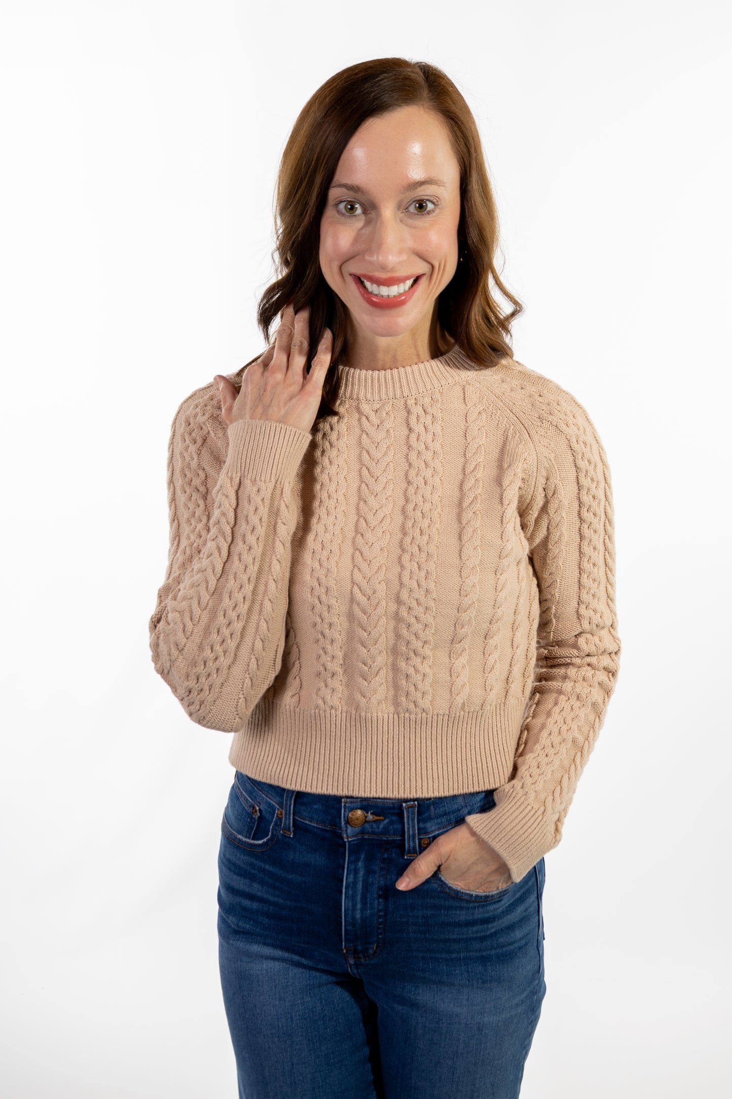 Woman wearing a beige cable knit sweater and blue jeans on a white background