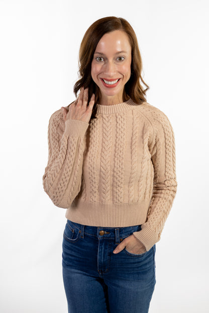Woman wearing a beige cable knit sweater and blue jeans on a white background
