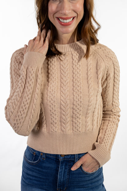Woman wearing a beige cable knit sweater and blue jeans on a white background