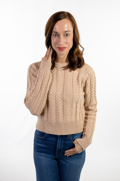 Woman wearing a beige cable knit sweater and blue jeans on a white background