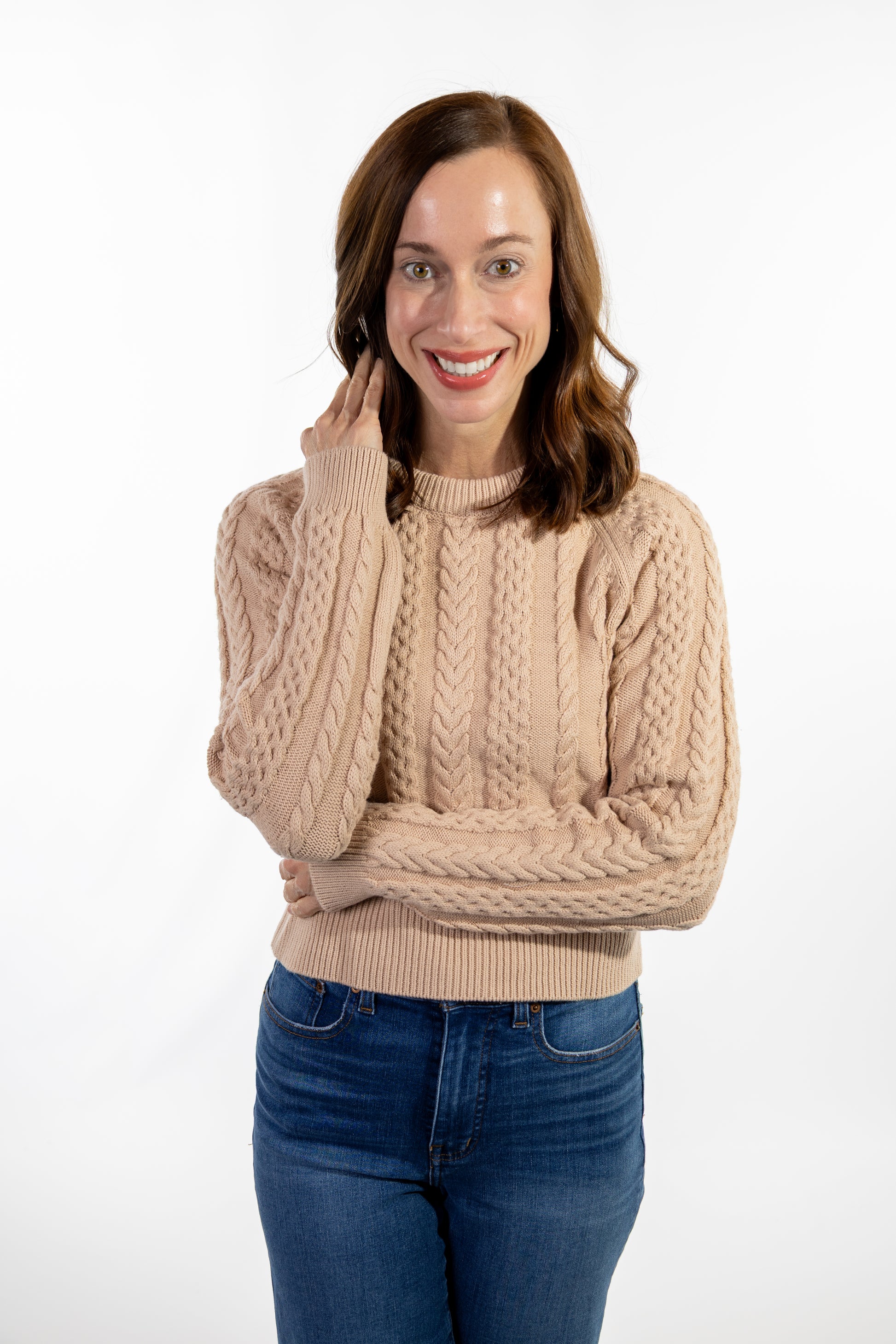 Woman wearing a beige cable knit sweater and blue jeans on a white background