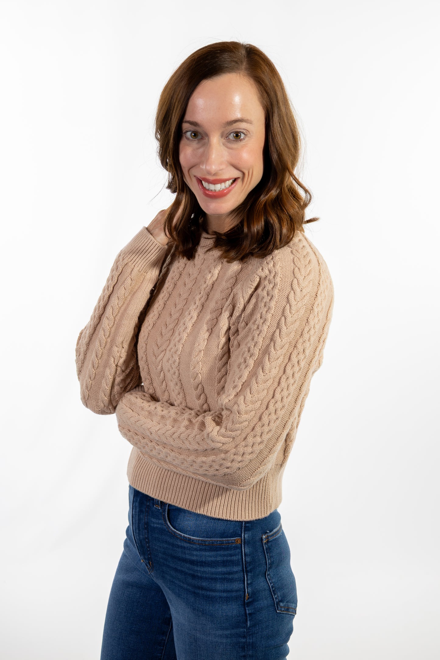 Woman wearing a beige cable knit sweater and blue jeans on a white background