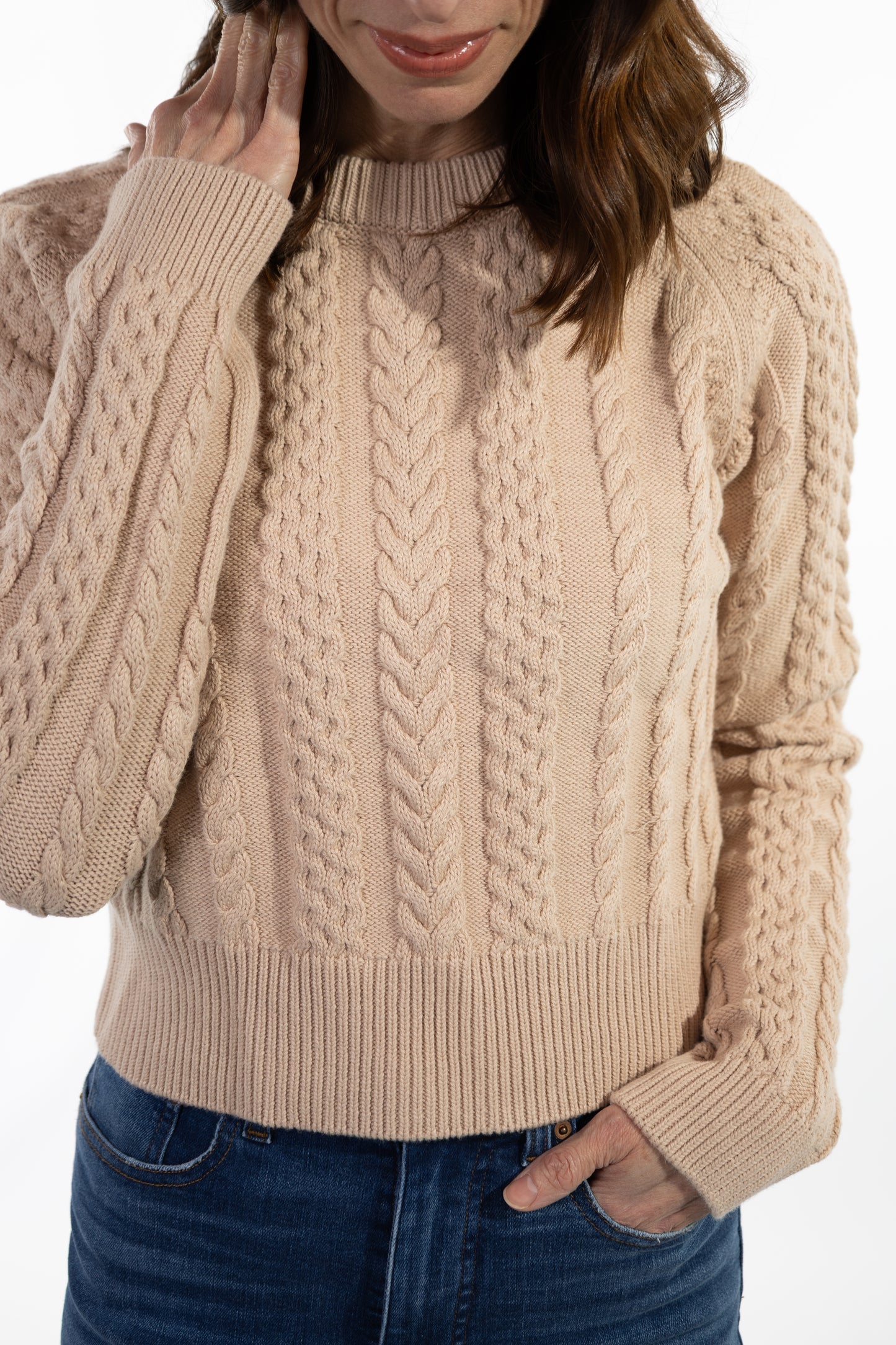 Person wearing a beige cable knit sweater with blue jeans on a white background