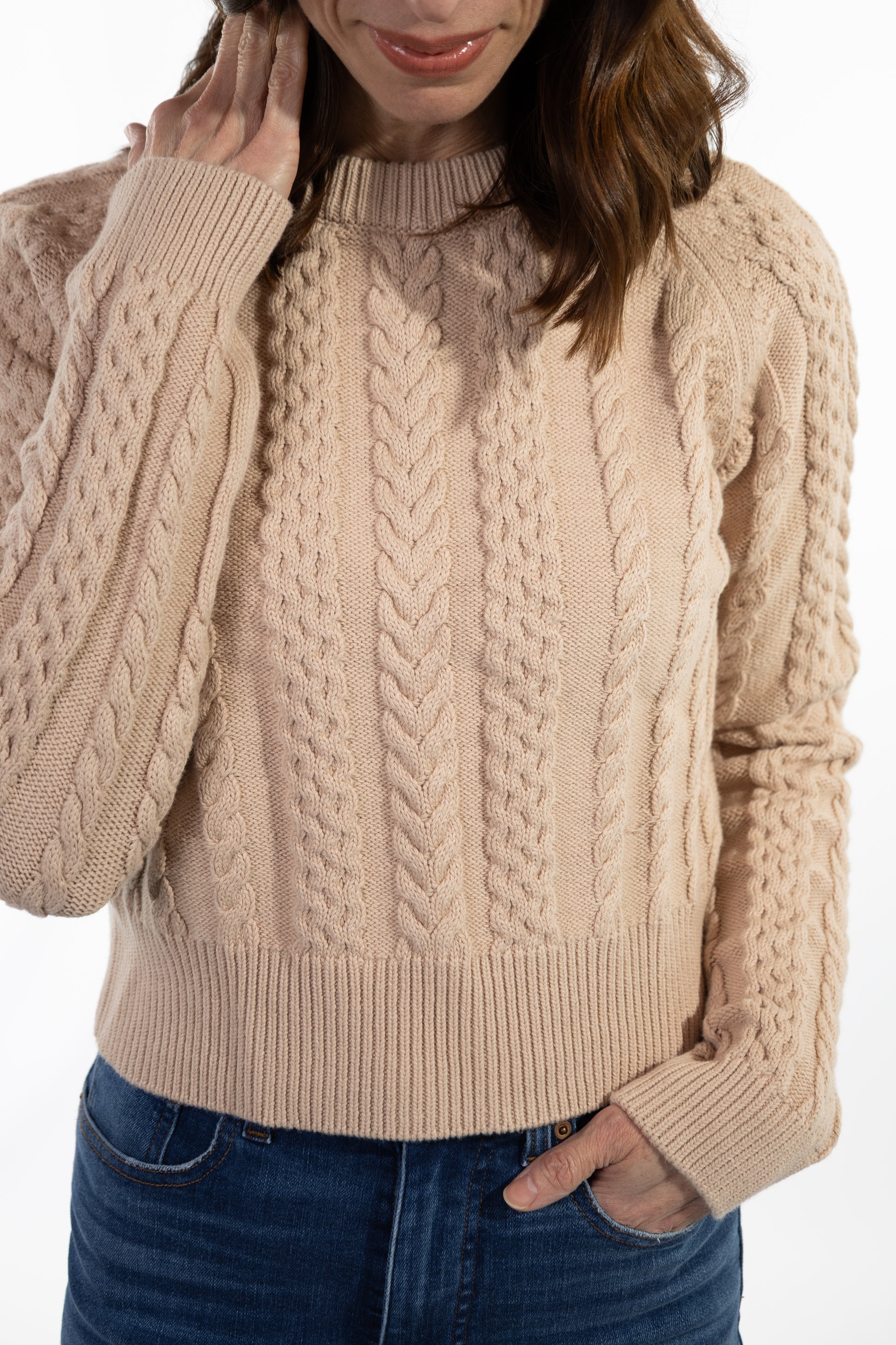 Person wearing a beige cable knit sweater with blue jeans on a white background