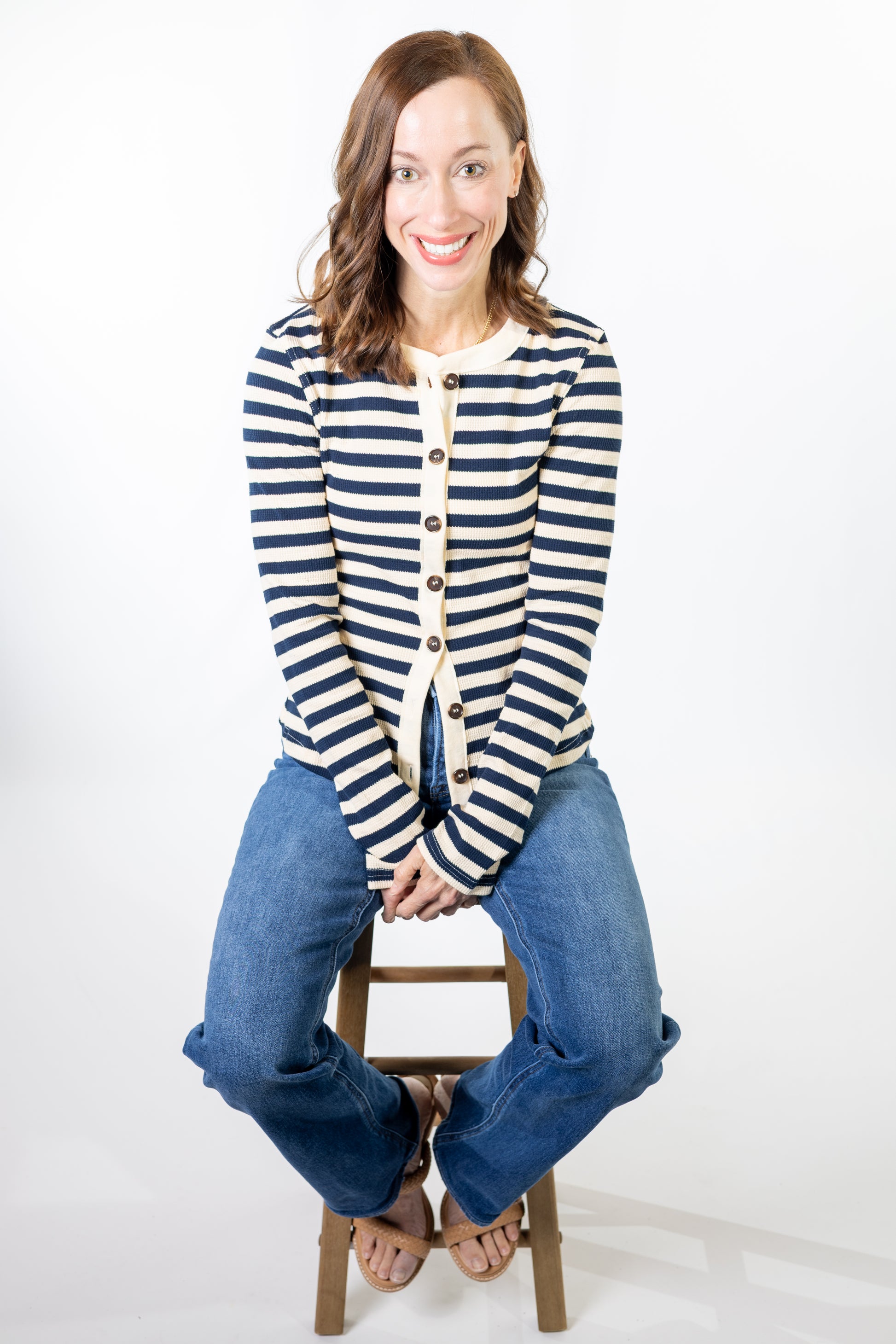 Woman wearing a striped cardigan and jeans sitting on a stool against a white background