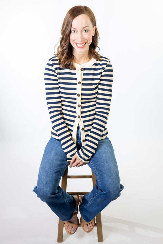 Woman wearing a striped cardigan and jeans sitting on a stool against a white background