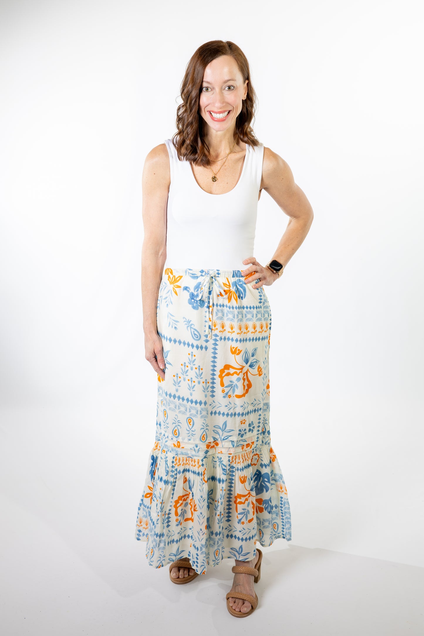 Woman wearing a white sleeveless top and a long, patterned blue, ivory and coral orange skirt on a white background