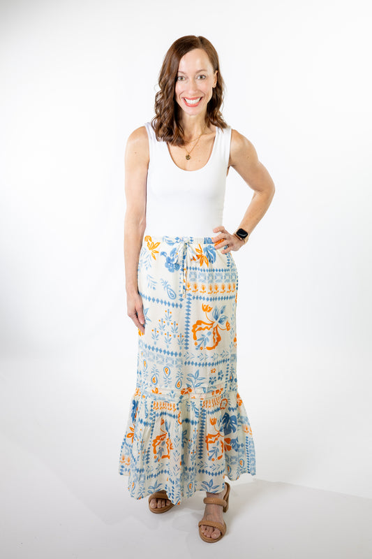 Woman wearing a white sleeveless top and a long, patterned blue, ivory and coral orange skirt on a white background