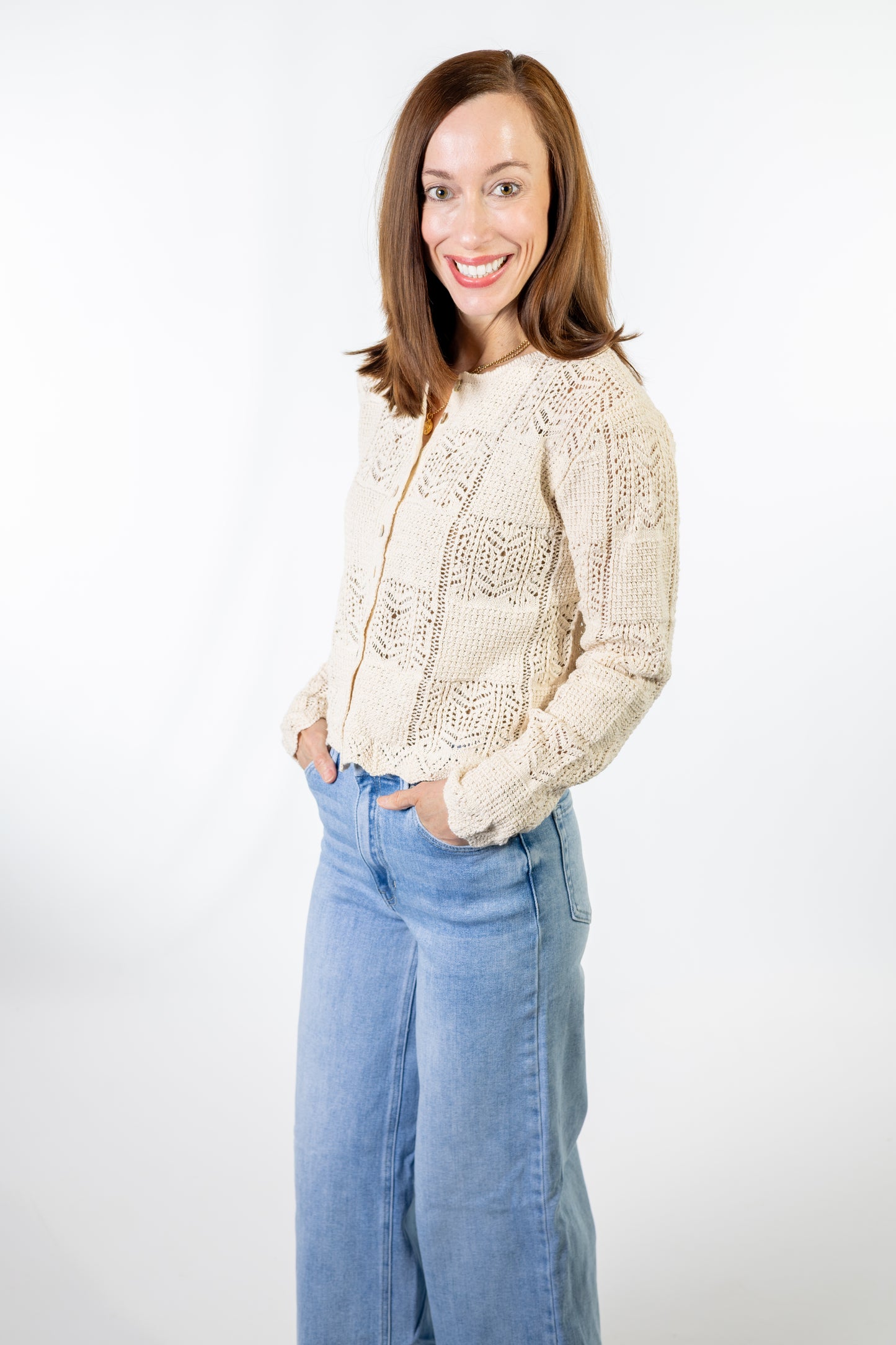 Woman wearing a beige lace crochet button up knit shirt and light blue jeans on a white background