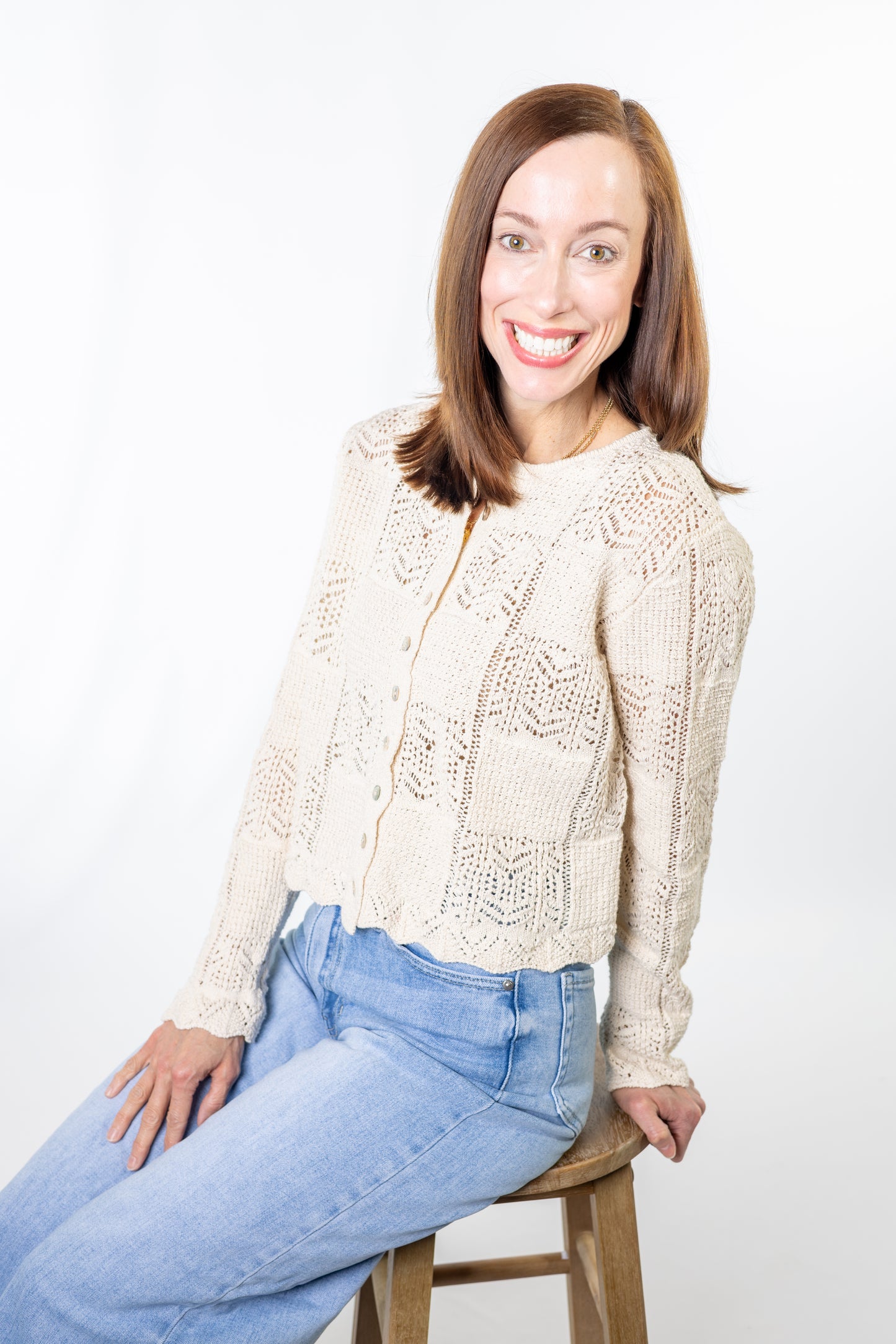 Woman wearing a beige cream crochet lace cardigan and blue jeans sitting on a wooden stool against a white background
