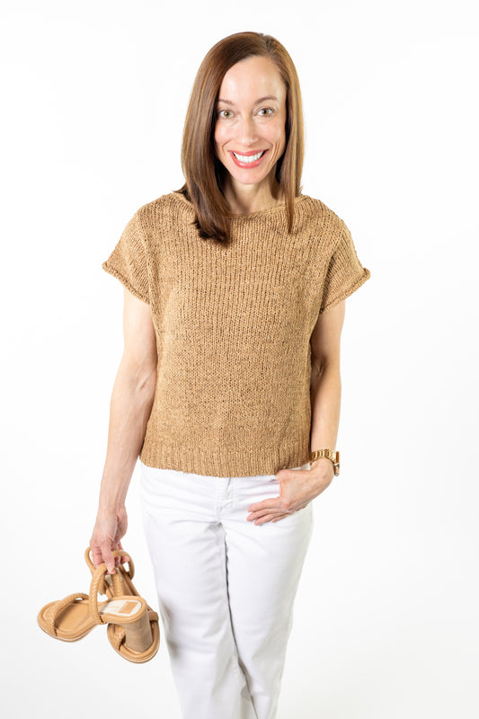 Woman wearing a beige sweater and white pants, holding sandals, on a white background