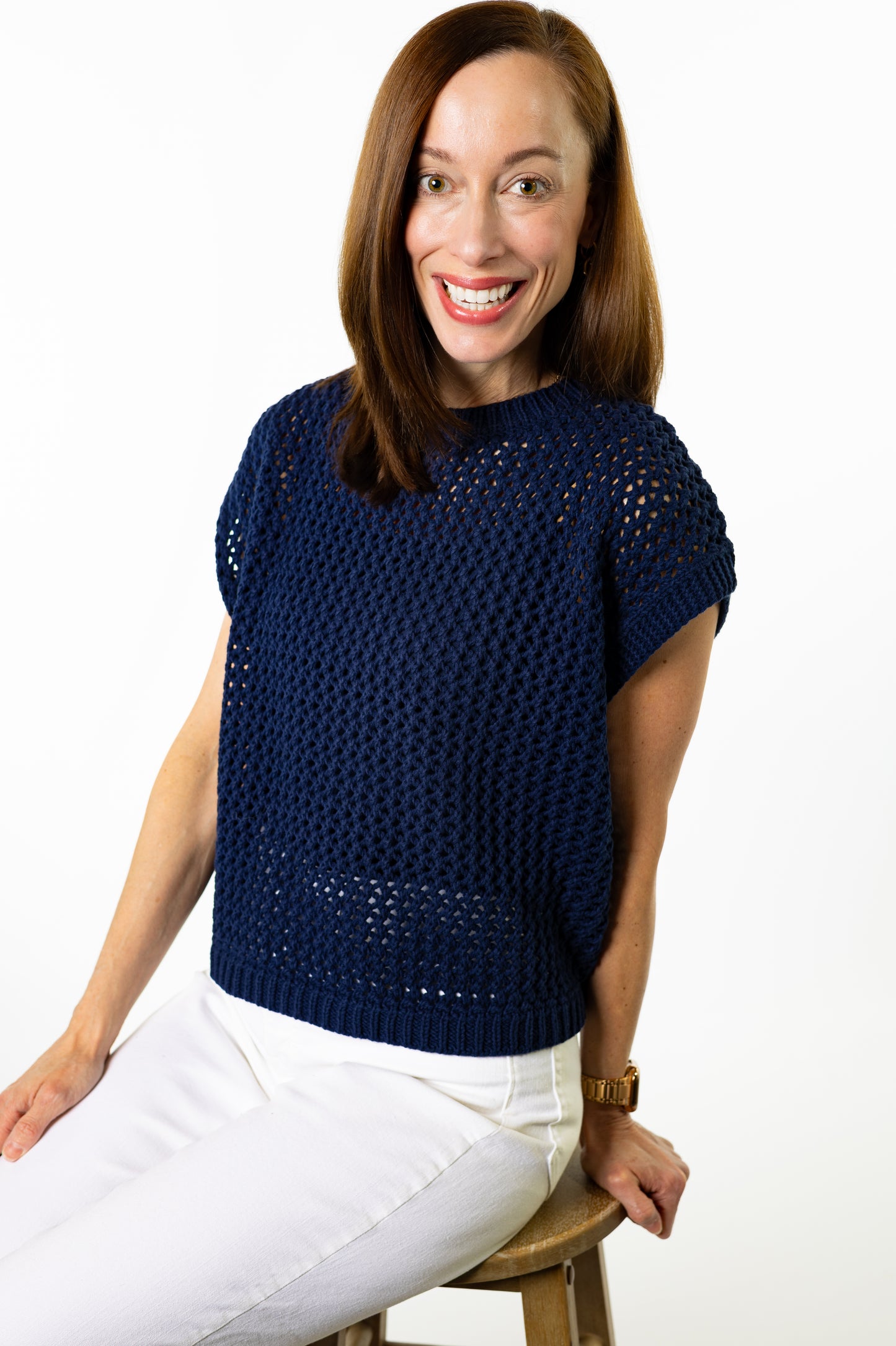 Woman wearing navy blue sweater with white pants and white background sitting on stool