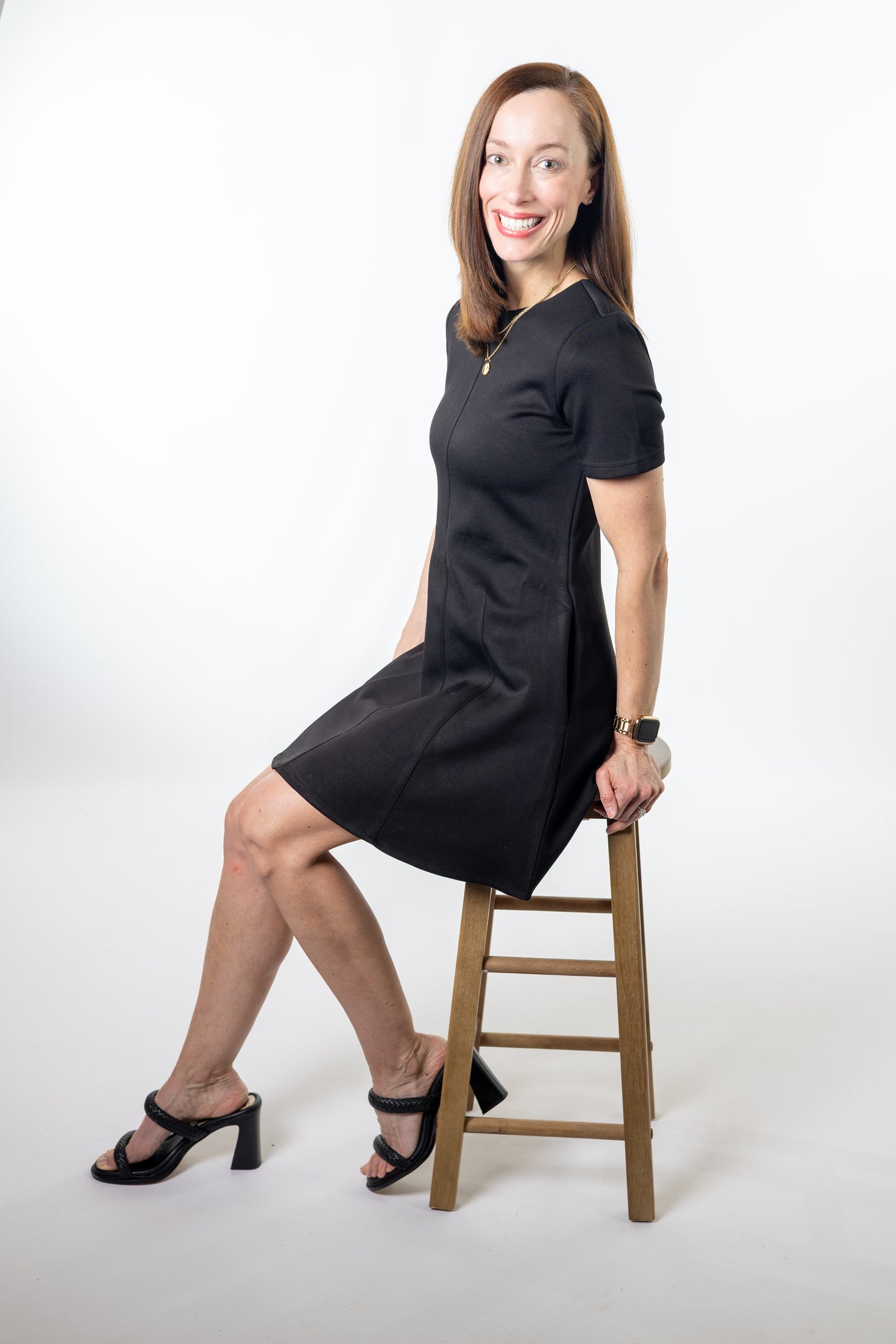 Woman in a black dress sitting on a wooden stool against a white background