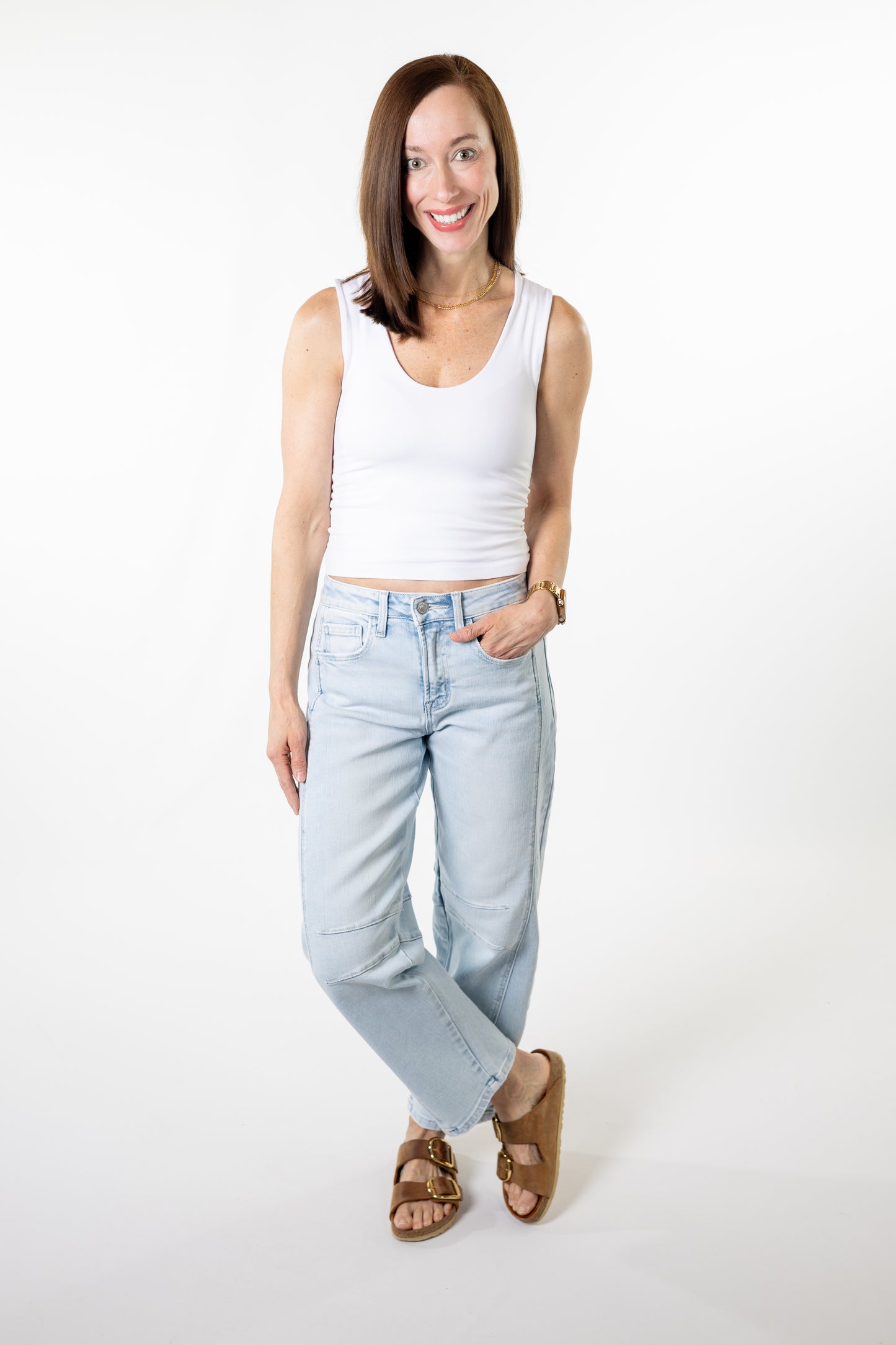 Woman wearing a white tank top and light blue high rise barrel jeans on a white background