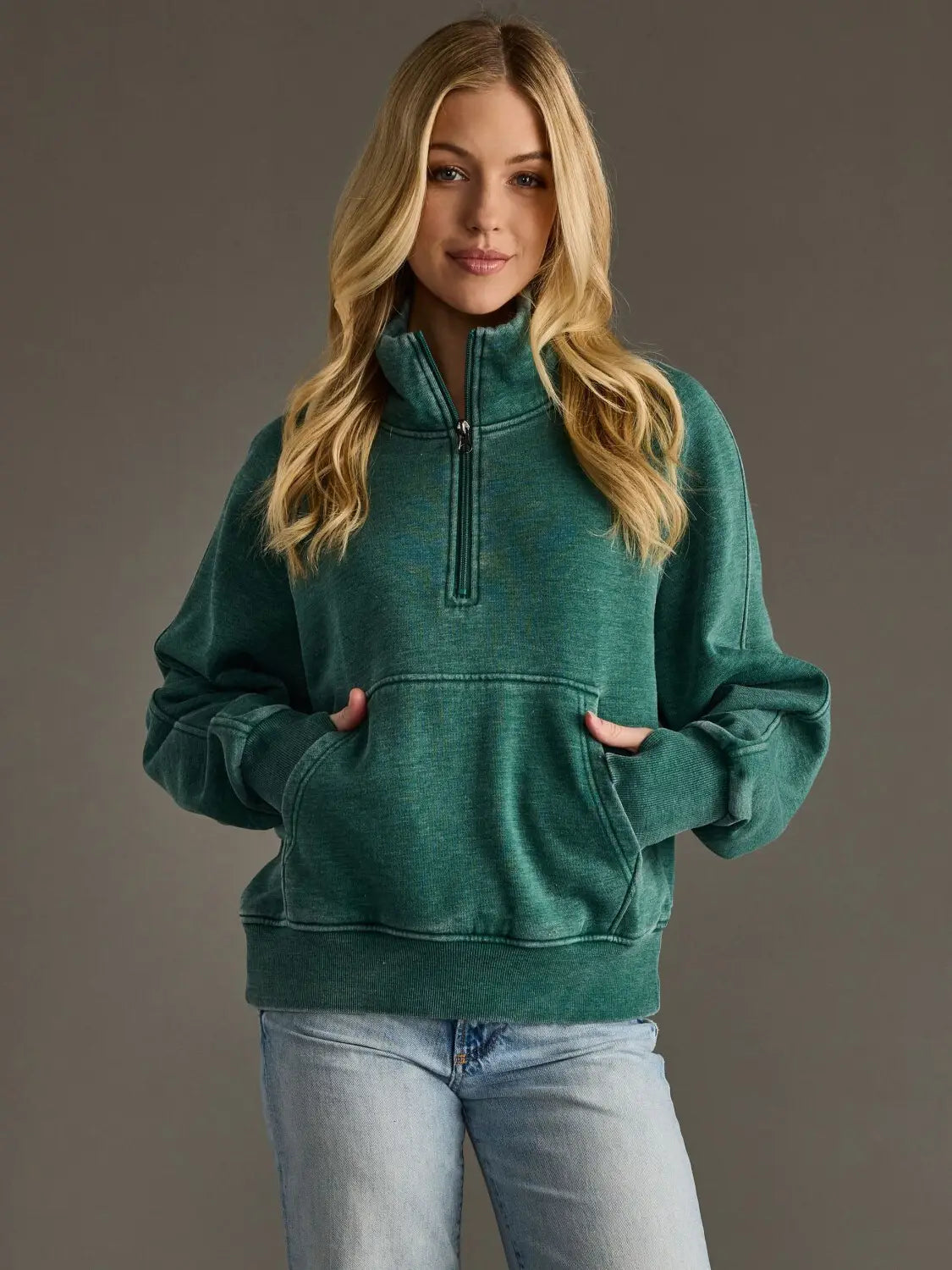 A woman with blonde hair wearing a dark green heathered quarter-zip sweatshirt and light blue jeans, standing against a solid dark gray studio background.